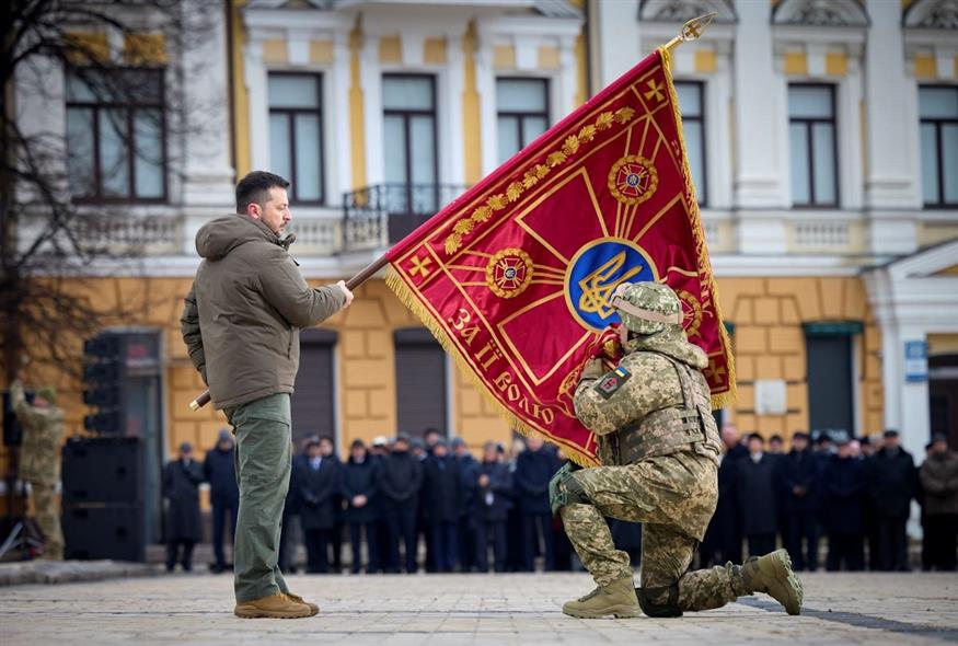 Ένας στρατιώτης φιλά μια σημαία με το σύμβολο του στρατού, την οποία κρατά ο Βολοντίμιρ Ζελένσκι (Ukrainian Presidential Press Office via AP)