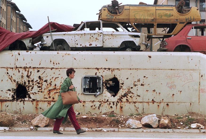 Μια Βόσνια γυναικά περνά μπροστά από ένα οδόφραγμα φτιαγμένο από λεωφορεία και αυτοκίνητα που έχουν υποστεί ζημιές από όλμους και σφαίρες στο Σαράγεβο, το 1994 (Jacqueline Arzt/AP)